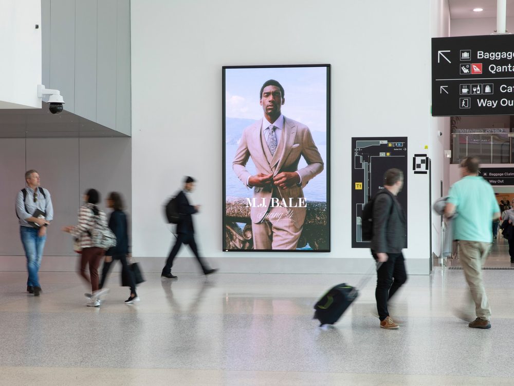 M.J. Bale campaign on a large digital screen in an airport terminal, with travellers walking past